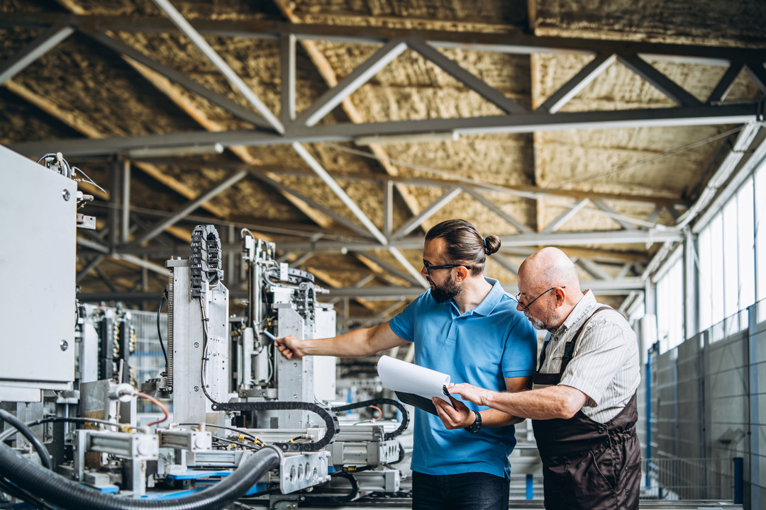 EPDM: um trabalhador jovem aponta para máquinas em funcionamento enquanto um colaborador idoso confere papéis em uma prancheta.