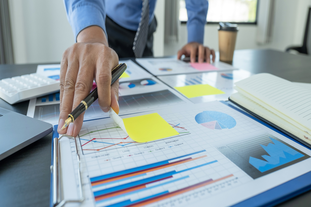Na foto, observamos um homem de negócios com uma caneta na mão analisando um relatório de atingimento de metas.